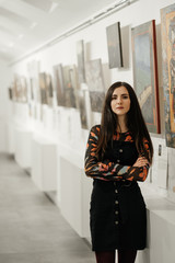 Young beautiful brunette girl standing in a gallery contemplating paintings artworks displayed on gallery walls. Art gallery.