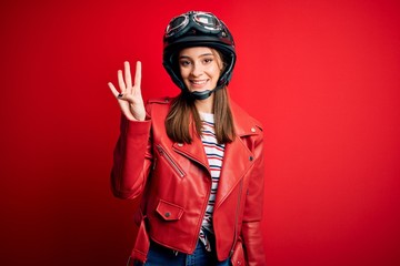 Young beautiful brunette motocyclist woman wearing motorcycle helmet and red jacket showing and pointing up with fingers number four while smiling confident and happy.