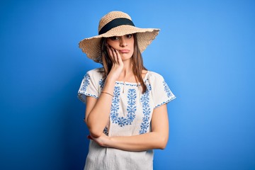 Young beautiful brunette woman wearing casual t-shirt and summer hat over blue background thinking looking tired and bored with depression problems with crossed arms.