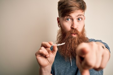 Irish redhead man with beard holding dental invisible aligner for tooth correction pointing with finger to the camera and to you, hand sign, positive and confident gesture from the front