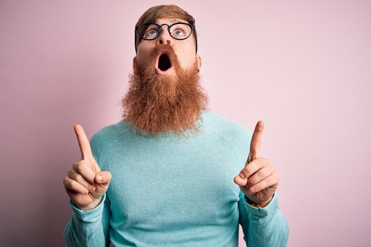 Handsome Irish redhead man with beard wearing glasses over pink isolated background amazed and surprised looking up and pointing with fingers and raised arms.