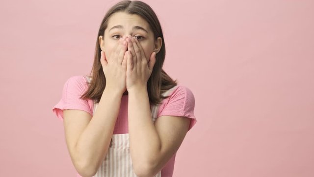 Concentrated pretty brunette woman in overalls looking at the camera then becoming shocked over pink background