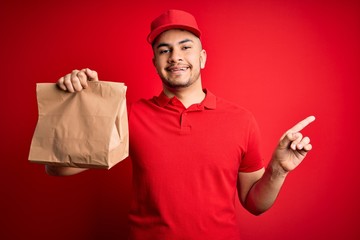 Young handsome delivery man holding paper bag with takeaway food over red background very happy pointing with hand and finger to the side