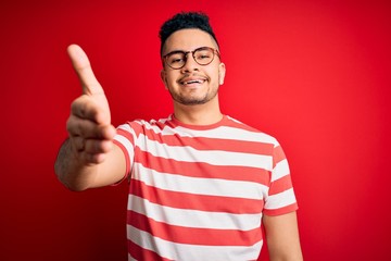 Naklejka premium Young handsome man wearing casual striped t-shirt and glasses over isolated red background smiling friendly offering handshake as greeting and welcoming. Successful business.