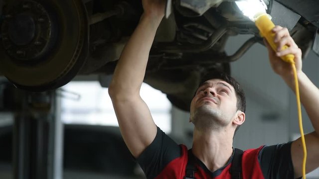A professional car mechanic works in a workshop, he checks the car raised up on the lift.
