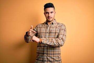 Young handsome man wearing casual shirt standing over isolated yellow background In hurry pointing...