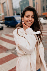 Fototapeta premium Outside portrait of attractive woman with dark hair wearing white coat walking on the street with charming smile in good day