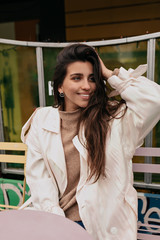 Close-up portrait of cheerful woman in white coat her hair on open air terrace. Photo of fashionable girl with dark hair smiling to camera.