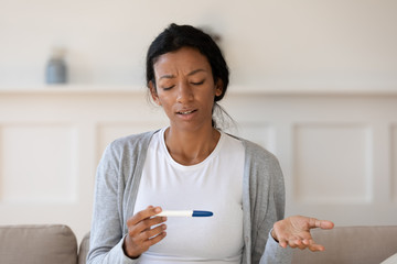 Upset young black girl hold pregnancy test in hand, unexpected or unwanted pregnancy, thinking about abortion. Frustrated by negative result, problem with infertility or health, maternity concept - Powered by Adobe
