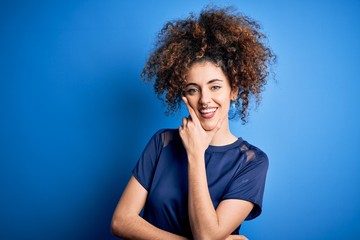 Young beautiful woman with curly hair and piercing wearing casual blue t-shirt looking confident at the camera smiling with crossed arms and hand raised on chin. Thinking positive.