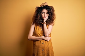 Beautiful brunette woman on vacation with curly hair and piercing wearing hat and dress thinking looking tired and bored with depression problems with crossed arms.