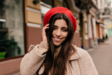 Carefree girl in elegant hat posing at camera on background of old city. Outdoor portrait of blissful long-haired woman in beige coat laughing while walking on the street