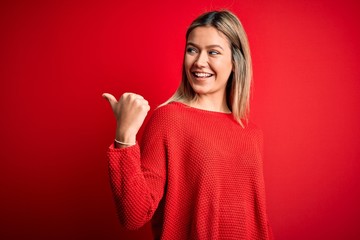 Young beautiful blonde woman wearing casual sweater over red isolated background smiling with happy face looking and pointing to the side with thumb up.