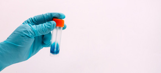 A hand in a blue latex glove holds a container for collecting feces. Plastic container for analysis in the doctors hand. White background.