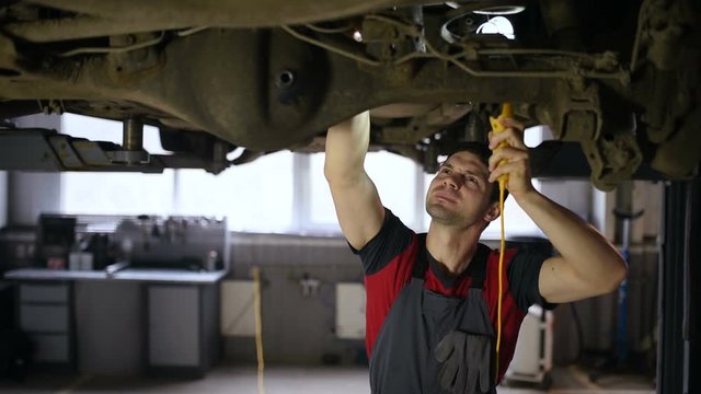 A professional car mechanic works in a workshop, he checks the car raised up on the lift.