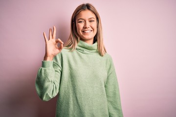 Young beautiful blonde woman wearing winter wool sweater over pink isolated background smiling positive doing ok sign with hand and fingers. Successful expression.