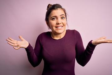 Beautiful young woman wearing casual bun hairstyle over pink isolated background clueless and confused expression with arms and hands raised. Doubt concept.