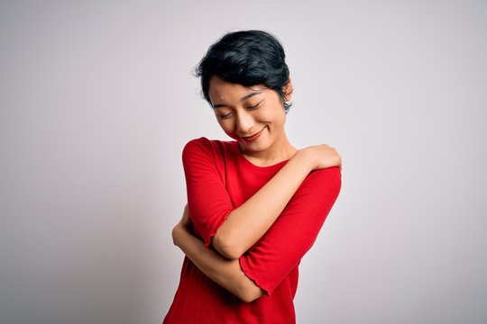 Young beautiful asian girl wearing casual red t-shirt standing over isolated white background Hugging oneself happy and positive, smiling confident. Self love and self care