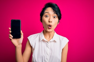 Young beautiful asian girl holding smartphone showing screen over isolated pink background scared...