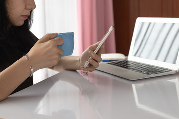 Fototapeta na wymiar Woman drinking and using smartphone with computer laptop and working at home for business, self-quarantine, staying home and social distancing in coronavirus or Covid-2019 outbreak situation concept
