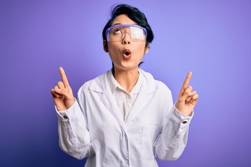 Young beautiful asian scientist girl wearing coat and glasses over purple background amazed and surprised looking up and pointing with fingers and raised arms.