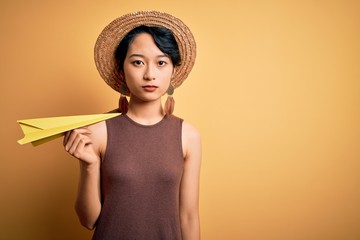 Young beautiful chinese woman on vacation wearing summer hat holding paper airplane with a confident expression on smart face thinking serious