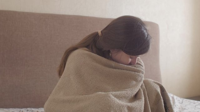 young woman trembling from cold wrapped in blanket sitting on the bed in bedroom interior, girl suffers from runny nose blowing in paper towel, concept of illness, allergy and health
