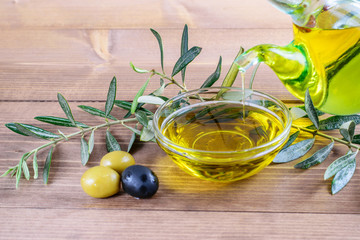 Olive oil dripping in the glass transparent bowl. Fresh olives, branches of olive tree on the wooden background.