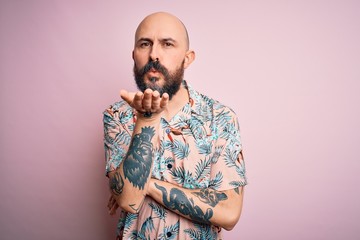 Handsome bald man with beard and tattoo wearing casual floral shirt over pink background looking at...