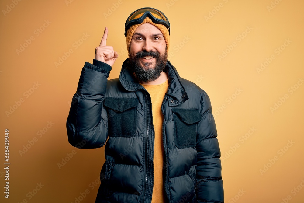 Poster handsome skier bald man with beard skiing wearing snow sportswear and ski goggles pointing finger up