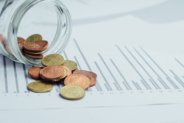 Glass jar with money, coins spilled from a piggy bank on graphic charts background. Little money or small salary. Break into savings, crisis, bankruptcy concept. 