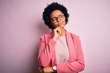 Young beautiful African American afro businesswoman with curly hair wearing pink jacket with hand on chin thinking about question, pensive expression. Smiling with thoughtful face. Doubt concept.