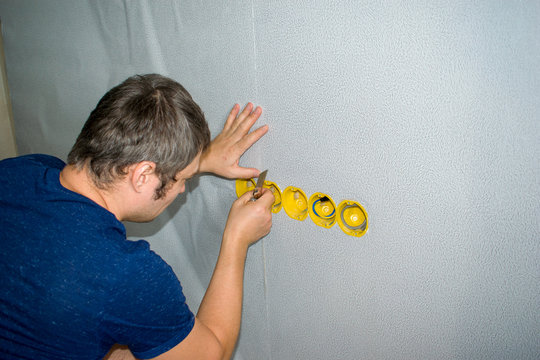 The man cuts the wallpaper on the walls in the places of the sockets with a knife.