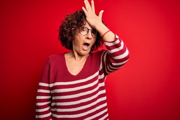 Middle age beautiful curly hair woman wearing casual striped sweater over red background surprised...