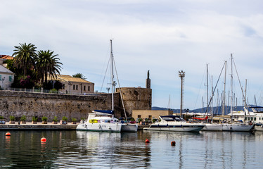 Fototapeta na wymiar Classic white yachts anchored in the ancient port of Alghero with palms, brick tower and wall. Sardinia, Italia. Beautiful view on the seafront.