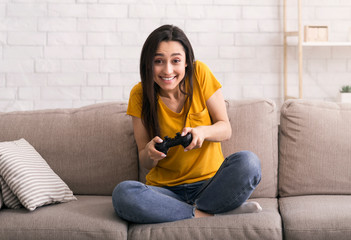 Fototapeta premium Quarantine pastimes. Joyful Latin girl with joystick playing video games on sofa at home