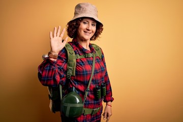 Fototapeta na wymiar Middle age curly hair hiker woman hiking wearing backpack and water canteen using binoculars Waiving saying hello happy and smiling, friendly welcome gesture