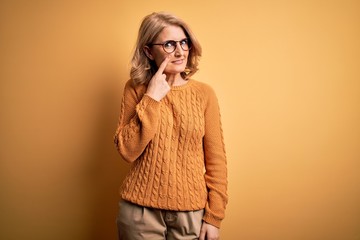 Middle age beautiful blonde woman wearing casual sweater and glasses over yellow background Pointing with hand finger to face and nose, smiling cheerful. Beauty concept