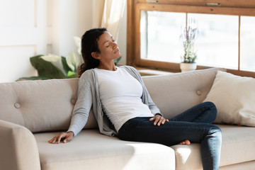 Obraz premium Black millennial woman sit and rest on comfortable sofa in cozy modern living room. Cute and serene woman with closed eyes take a nap on couch, enjoy calm and silence. Break at home. Feel good