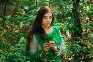 Attractive girl in a green medieval dress sits in the bushes of fern. Village woman herbalist collects herbs for potion in the forest.