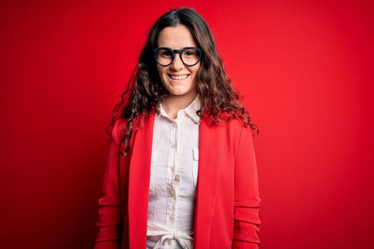 Young beautiful woman with curly hair wearing jacket and glasses over red background with a happy and cool smile on face. Lucky person.