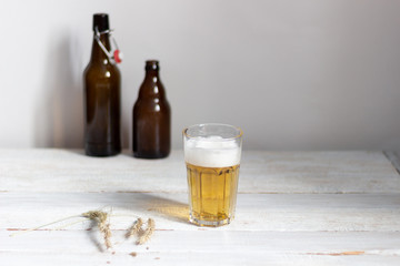 Glass of homemade beer on white wooden background with barley spikes, bottles