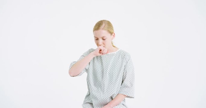Young female medical patient in hospital night dress caughing a lot as being sick. Beautiful ill woman having caugh spasm on white wall background. Coronavirus symptoms concept. Virus progressing.