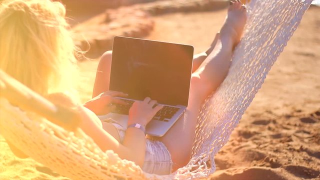 Beautiful girl lies in a hammock and works at a laptop on the beach.