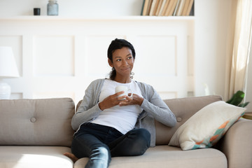 Young millennial calm african american girl relaxing and sit on comfortable sofa in living room, relaxed weekend at home, woman drinking tea or coffee and dreaming or thinking