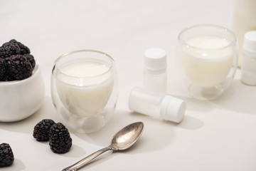 High angle view of glasses of yogurt, teaspoon, containers with starter cultures and sugar bowl with blackberries on white