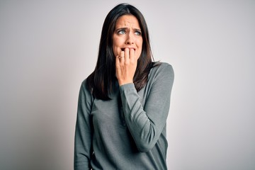 Young brunette woman with blue eyes wearing casual green sweater over white background looking stressed and nervous with hands on mouth biting nails. Anxiety problem.