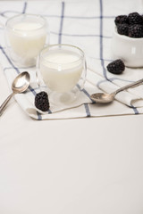 Selective focus of glasses of homemade yogurt, teaspoons and sugar bowl with blackberries on cloth on white