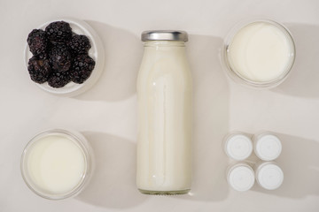 Top view of bottle and glasses of homemade yogurt, containers with starter cultures and sugar bowl with blackberries on white