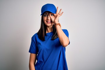Young delivery woman with blue eyes wearing cap standing over blue background doing ok gesture with hand smiling, eye looking through fingers with happy face.